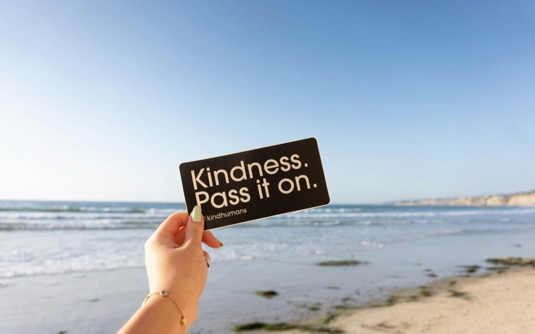 A notice held by a hand against the backdrop of the seak. It reads: Kindness. Pass It On.