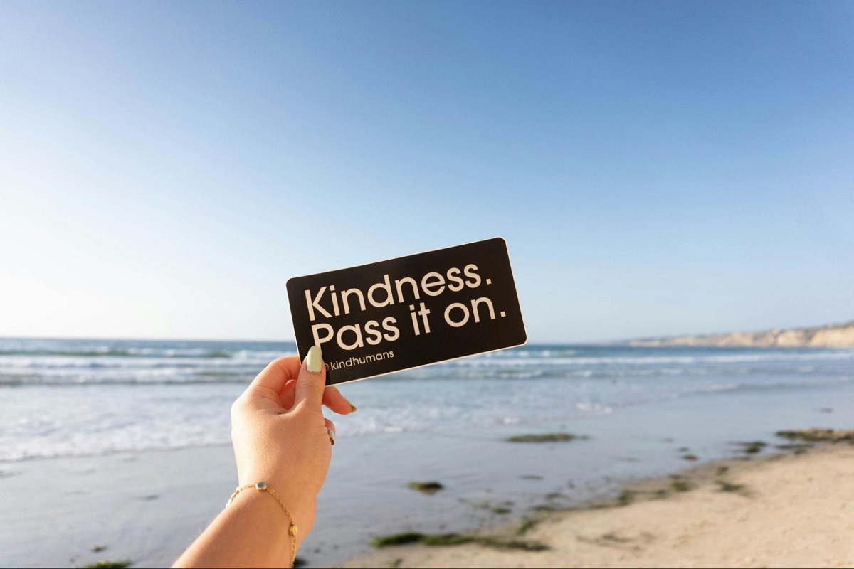 A notice held by a hand against the backdrop of the seak. It reads: Kindness. Pass It On.